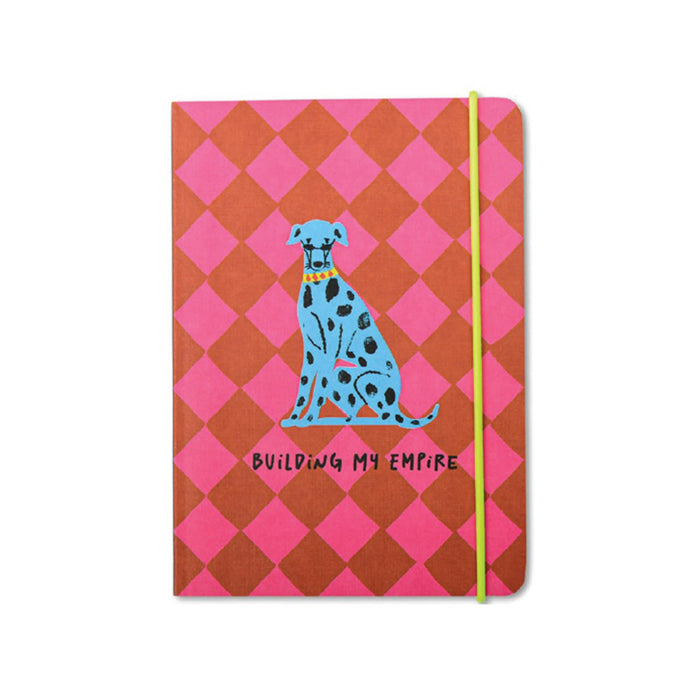 A softcover notebook with a pink and red diamond pattern, featuring a blue Dalmatian illustration and the text 'BUILDING MY EMPIRE' on the front cover, secured with a lime green elastic band.