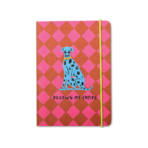 A softcover notebook with a pink and red diamond pattern, featuring a blue Dalmatian illustration and the text 'BUILDING MY EMPIRE' on the front cover, secured with a lime green elastic band.