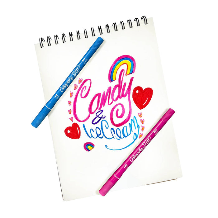 A white spiral-bound notepad with colorful hand-lettered 'Candy & Ice Cream' artwork, shown on a white background with two markers placed on it: one blue and one pink. The markers are branded Calligraphy Duo.