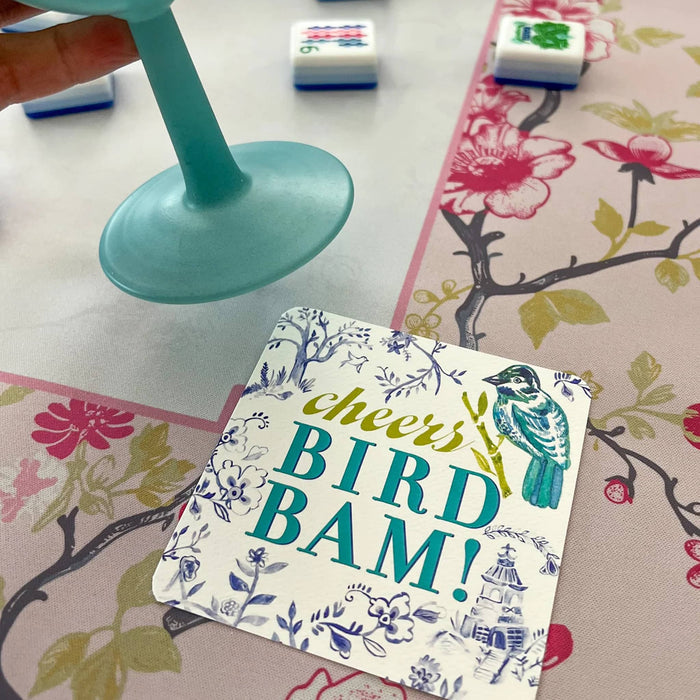A square printed coaster with the phrase “Cheers BIRD BAM!” and a bird illustration, placed on a floral table mat. A hand is holding a teal glass stem, and Mahjong tiles are visible in the background.