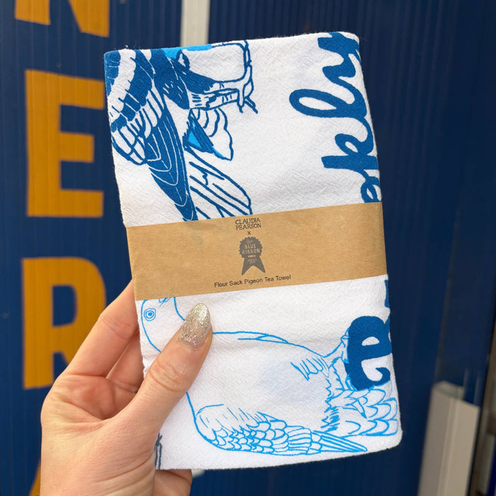 A hand holds a folded white tea towel with blue pigeon illustrations and 'brooklyn,' wrapped in a brown paper band that reads 'flour sack pigeon tea towel.' The background is a dark blue corrugated metal wall with yellow lettering.