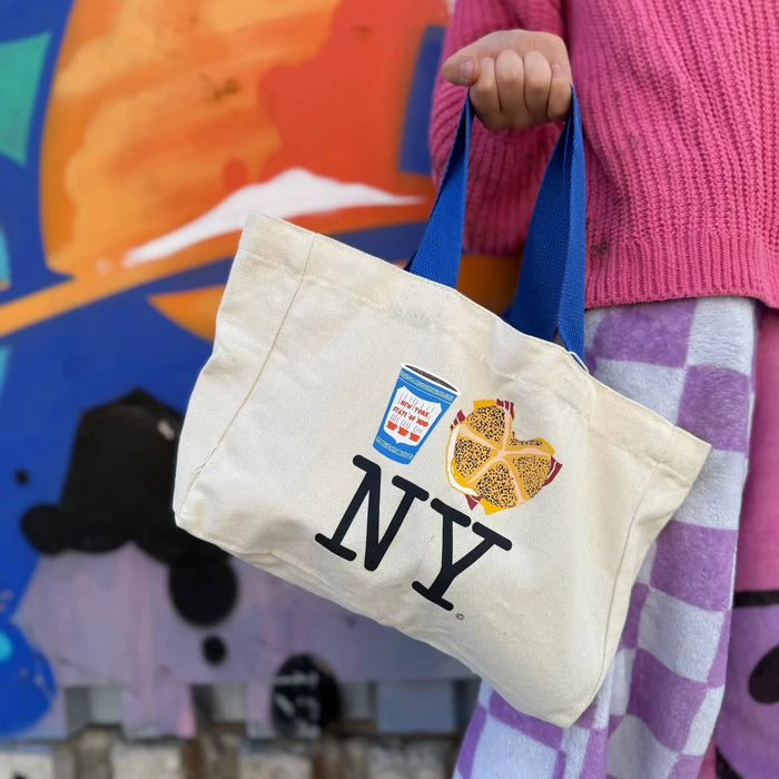 Person holding a mini tote bag with 'NY' and icons of a greek coffee cup and a bacon, egg, and cheese on a roll sandwich, against a colorful background.