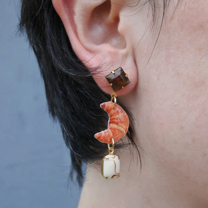 Close up of an ear with a drop earring with a croissant charm and vintage components, on a blue background.