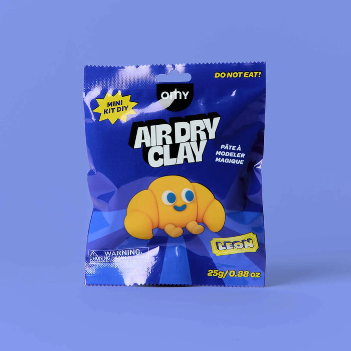 A blue bag of OMY Air Dry Clay mini kit, featuring a yellow croissant-shaped clay figure on the front, set against a solid periwinkle blue background.