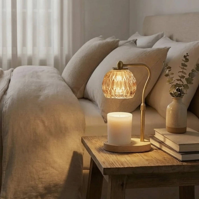 A warm, cozy bedroom scene featuring a lit candle warmer table lamp with a faceted glass shade and gold-toned metal frame, styled on a wooden bedside table next to a bed with beige linens and pillows.