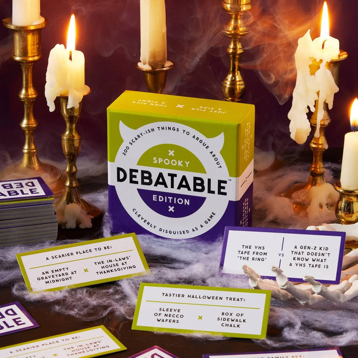 The Debatable Edition Spooky card game box is displayed on a dark, foggy surface surrounded by burning white candles in gold holders. Several sample cards are fanned out in front of the box, showing various Halloween-themed debate topics.