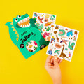 Children's tattoo sheets with dinosaur designs, with green dino-shaped packaging, on a yellow background.