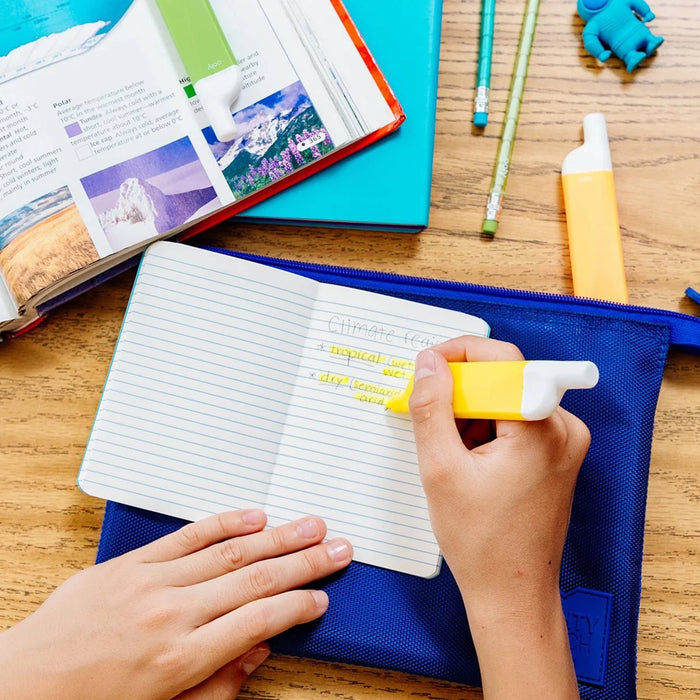 Person using a yellow highlighter on a ruled notebook with books and stationery items on a wooden surface.