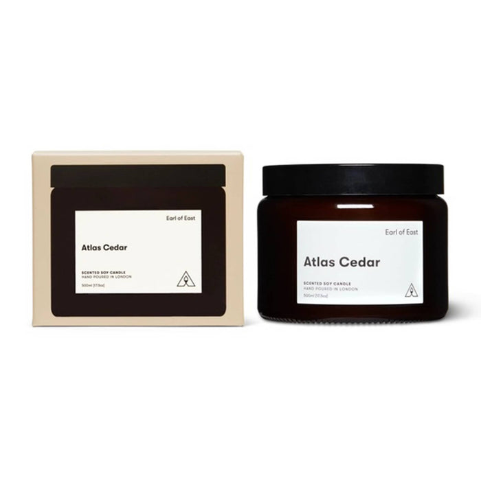 A scented soy candle in a dark amber glass jar with a black lid, shown next to its cream-colored cardboard packaging box. Both items feature a minimalist white label with the scent name 'Atlas Cedar' and the brand 'Earl of East'.