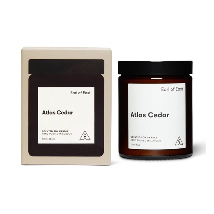A scented soy candle in a dark amber glass jar with a black lid, shown next to its cream-colored cardboard packaging box. Both items feature a minimalist white label with the scent name 'Atlas Cedar' and the brand 'Earl of East'.