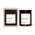 A scented soy candle in a dark amber glass jar with a black lid, shown next to its cream-colored cardboard packaging box. Both items feature a minimalist white label with the scent name 'Atlas Cedar' and the brand 'Earl of East'.