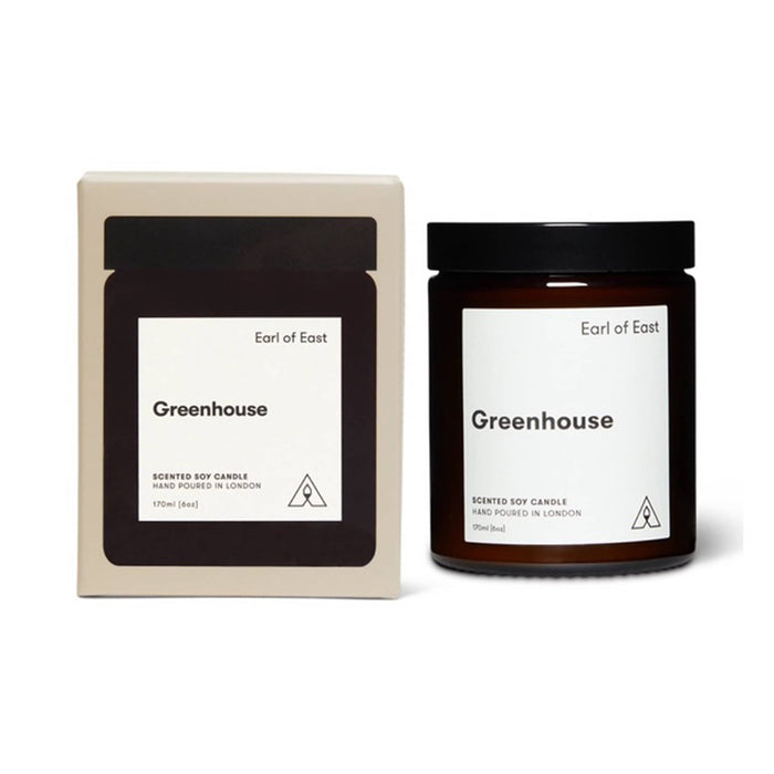 A scented soy candle in a dark amber glass jar with a black lid, shown next to its cream-colored cardboard packaging box. Both items feature a minimalist white label with the scent name 'Greenhouse' and the brand 'Earl of East'.