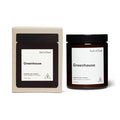 A scented soy candle in a dark amber glass jar with a black lid, shown next to its cream-colored cardboard packaging box. Both items feature a minimalist white label with the scent name 'Greenhouse' and the brand 'Earl of East'.