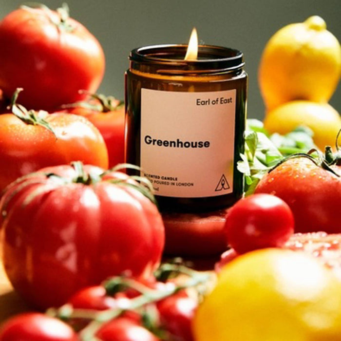A lit amber glass candle jar labeled 'Greenhouse' by Earl of East, surrounded by fresh tomatoes and lemons in a styled product photo.