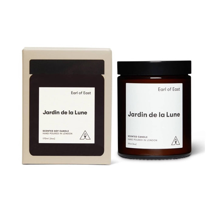 A scented soy candle in a dark amber glass jar with a black lid, shown next to its cream-colored cardboard packaging box. Both items feature a minimalist white label with the scent name 'Jardin de la Lune' and the brand 'Earl of East'.