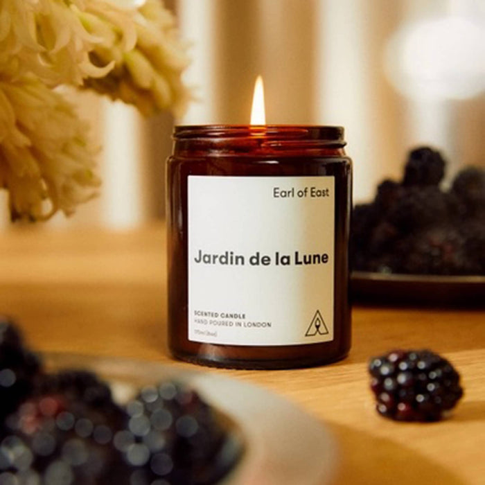 A lit scented candle in an amber glass jar sits on a wooden table, with a white label reading 'Earl of East' and 'Jardin de la Lune'. Blackberries and soft-focus flowers are visible around it in a warm, cozy setting.