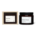 A scented soy candle in a dark amber glass jar with a black lid, shown next to its cream-colored cardboard packaging box. Both items feature a minimalist white label with the scent name 'Smoke & Musk' and the brand 'Earl of East'.