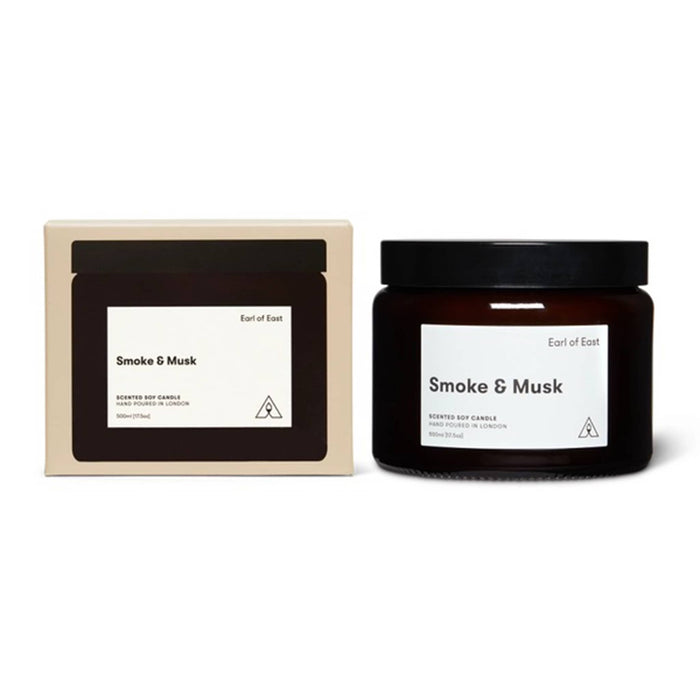A scented soy candle in a dark amber glass jar with a black lid, shown next to its cream-colored cardboard packaging box. Both items feature a minimalist white label with the scent name 'Smoke & Musk' and the brand 'Earl of East'.
