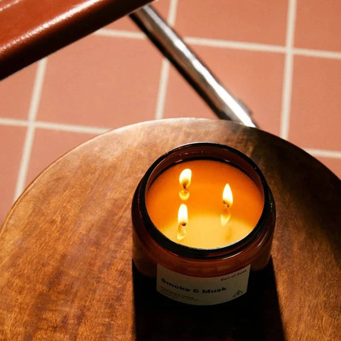 A lit amber glass candle with three wicks sits on a round wooden table, with terracotta tile flooring and a chair leg visible in the background. The label reads 'Smoke & Musk'.