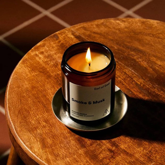 A lit amber glass candle jar labeled 'Smoke & Musk' by Earl of East, sitting on a silver saucer atop a round wooden stool in warm indoor lighting.