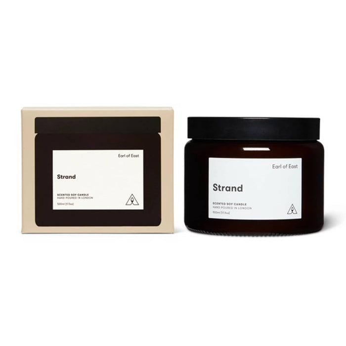 A scented soy candle in a dark amber glass jar with a black lid, shown next to its cream-colored cardboard packaging box. Both items feature a minimalist white label with the scent name 'Strand' and the brand 'Earl of East'.