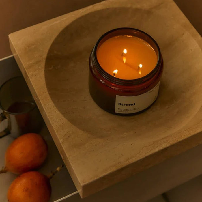 A lit amber glass jar candle with three wicks sits on a beige stone tray, with two oranges in a drawer beside it. The candle label reads 'Strand'.