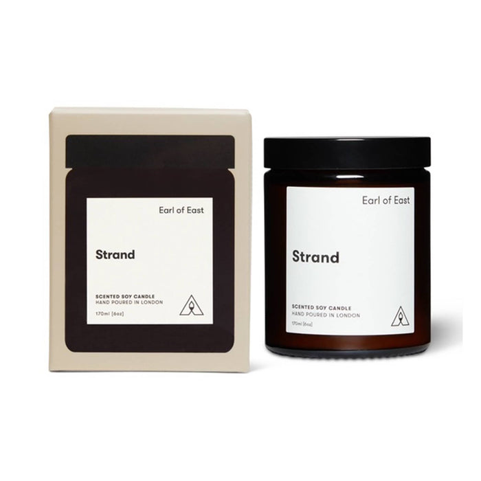 A scented soy candle in a dark amber glass jar with a black lid, shown next to its cream-colored cardboard packaging box. Both items feature a minimalist white label with the scent name 'Strand' and the brand 'Earl of East'.