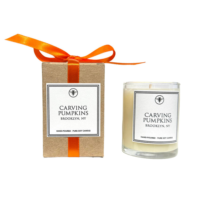 A hand-poured soy candle in a clear glass votive next to its kraft paper packaging box, both featuring white labels with black text that reads 'CARVING PUMPKINS BROOKLYN, NY' and tied with an orange ribbon.