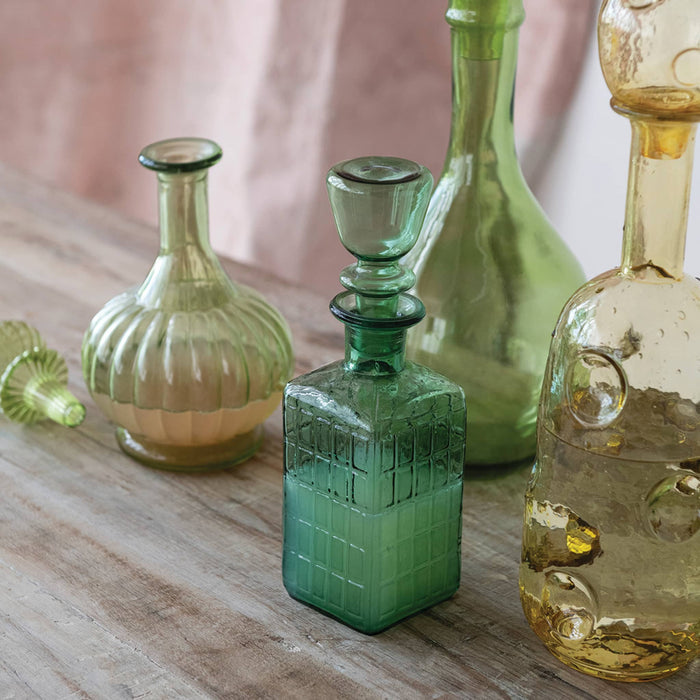 A collection of vintage green and amber glass decanters and bottles arranged on a rustic wooden table with a soft pink fabric backdrop.