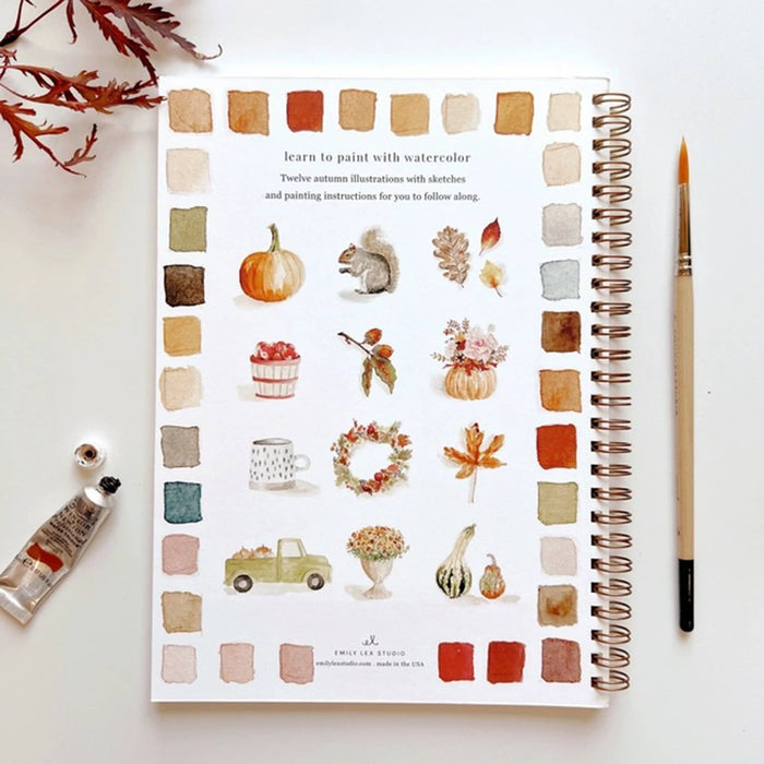 Autumn-themed watercolor workbook with color swatches and fall illustrations, surrounded by art supplies on a white background.