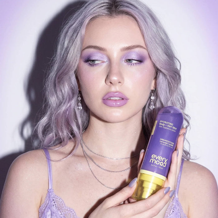 Woman with purple hair and makeup holding a bottle of Everymood Dreamcatcher scented hydrating body and hair mist, against a light purple  background.