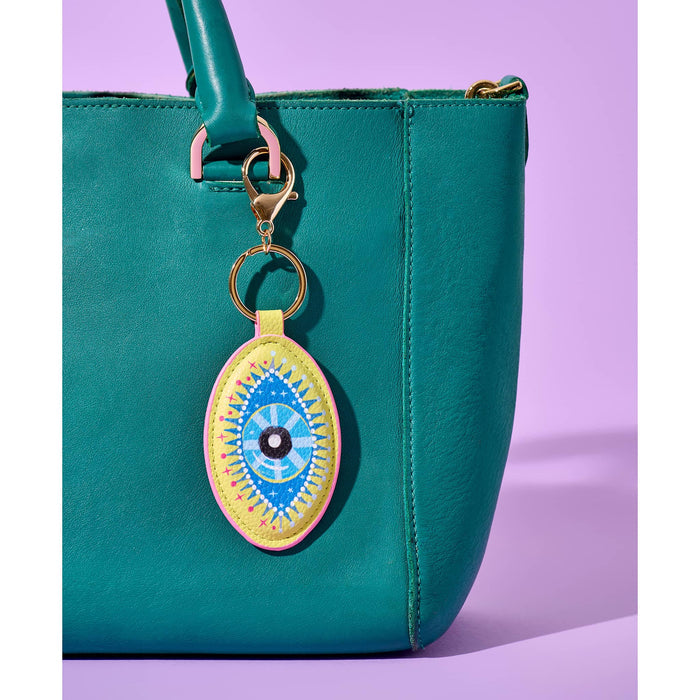 Green leather handbag with a yellow, blue, and pink leatherette bag charm attached to the handle, on a purple background.