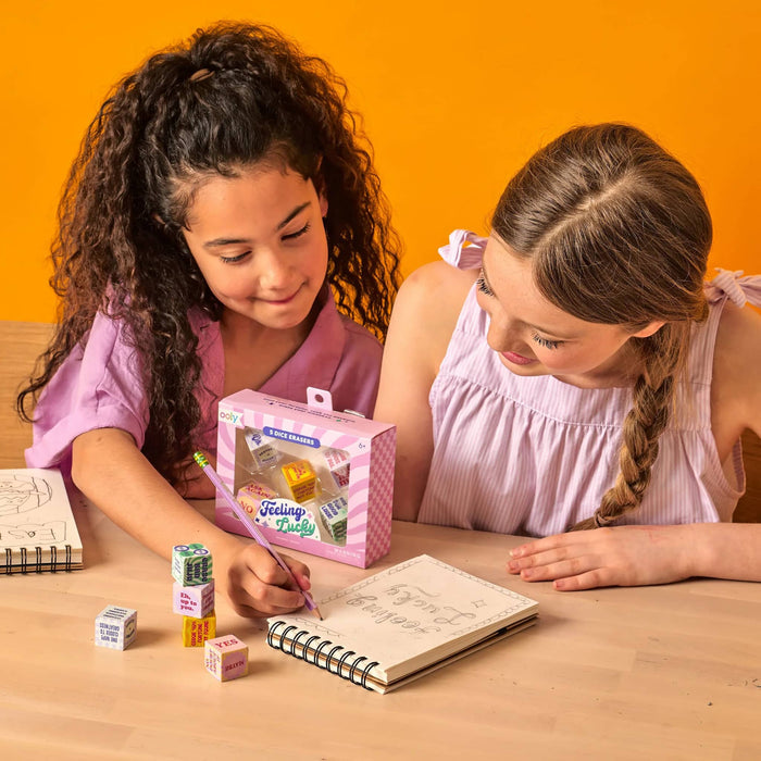 2 girls drawing in a spiral notebook with a package of dice-shaped erasers with a stack of them outside on a wood surface with an orange background.