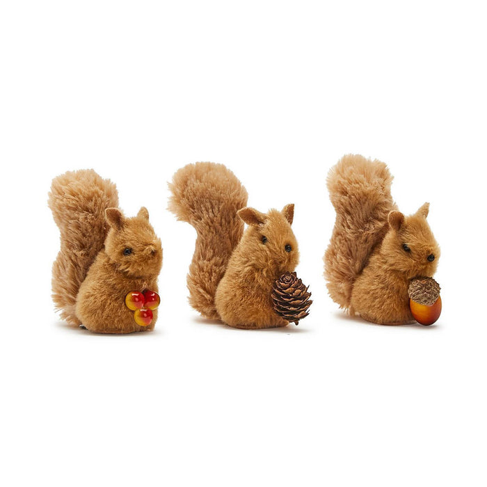 Three small brown plush squirrel figurines arranged side by side on an angle on a plain white background. The left squirrel holds red and yellow berries, the middle squirrel holds a pinecone, and the right squirrel holds an acorn.