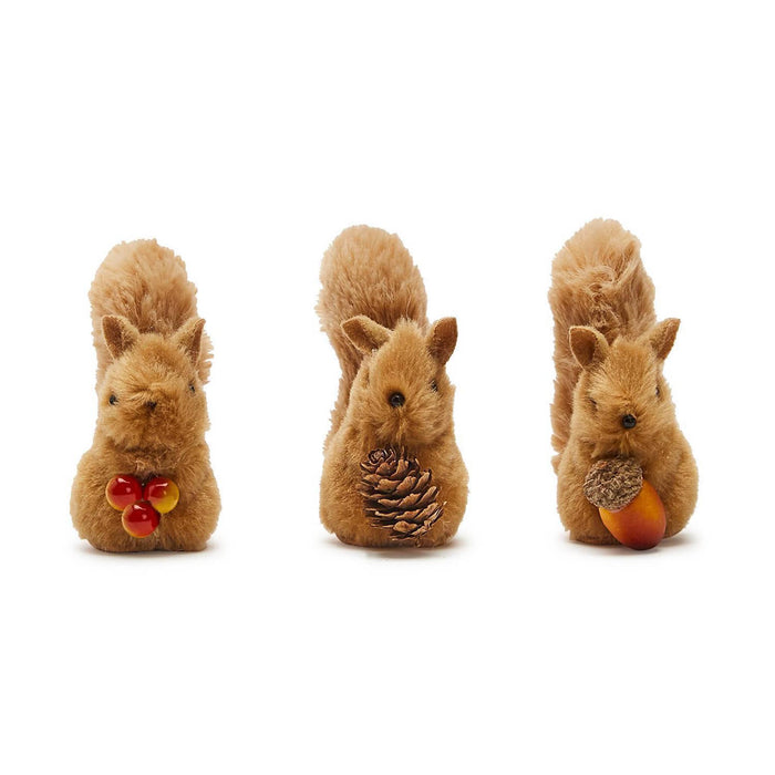Three small brown plush squirrel figurines arranged side by side, front view, on a plain white background. The left squirrel holds red and yellow berries, the middle squirrel holds a pinecone, and the right squirrel holds an acorn.