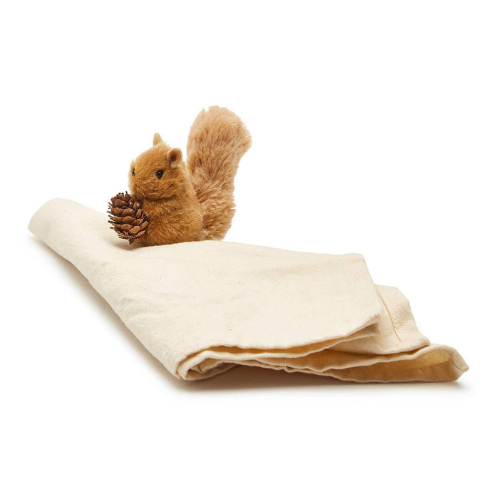 A plush brown squirrel figure holding a pinecone, posed on top of a folded cream-colored fabric napkin against a white background.