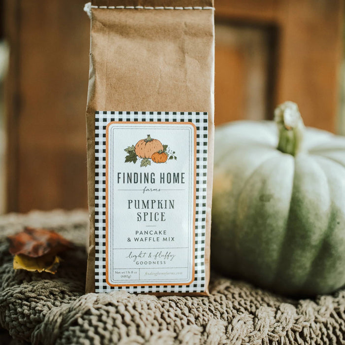 A bag of Finding Home Farms Pumpkin Spice Pancake & Waffle Mix sits on a knitted blanket with a rustic wooden background and a white pumpkin in the background.