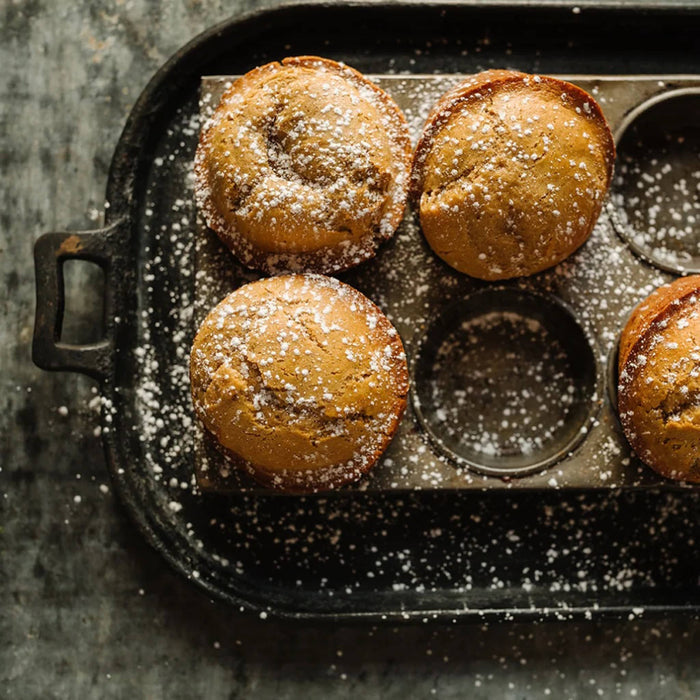 Four golden-brown muffins dusted with powdered sugar, arranged in a dark metal muffin tin on a rustic dark surface.