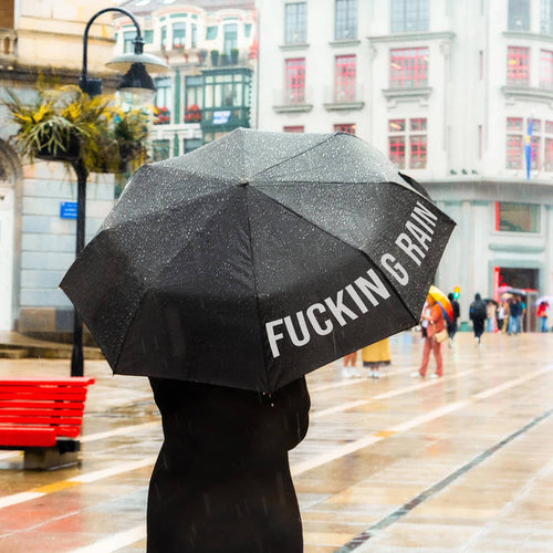 Person holding a black umbrella with 'FUCKING RAIN' text in an urban setting