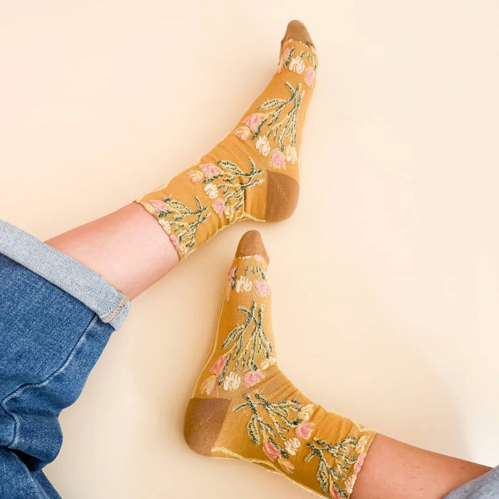 A pair of mustard yellow crew socks with a pink and green floral pattern, worn on feet with blue jeans rolled up against a solid beige background.