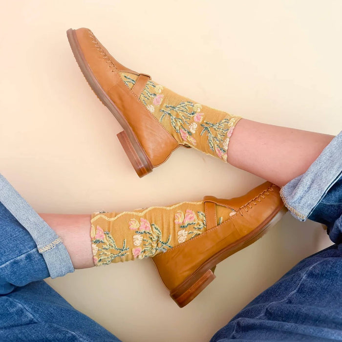 A person wearing tan leather loafers with floral embroidered socks, shown from the knees down against a light beige background.