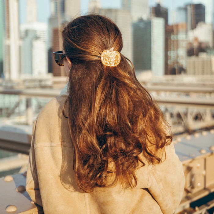 A woman seen from behind wearing a pumpkin hair claw clip with an intricate filigree pattern, holding her brown hair in a half-up style. She is wearing a beige jacket and sunglasses, with a blurred city skyline in the background.