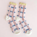 A pair of sheer white socks with pink and blue striped patterns, featuring pale yellow ribbed cuffs and white heels, laid flat on a light pink background.