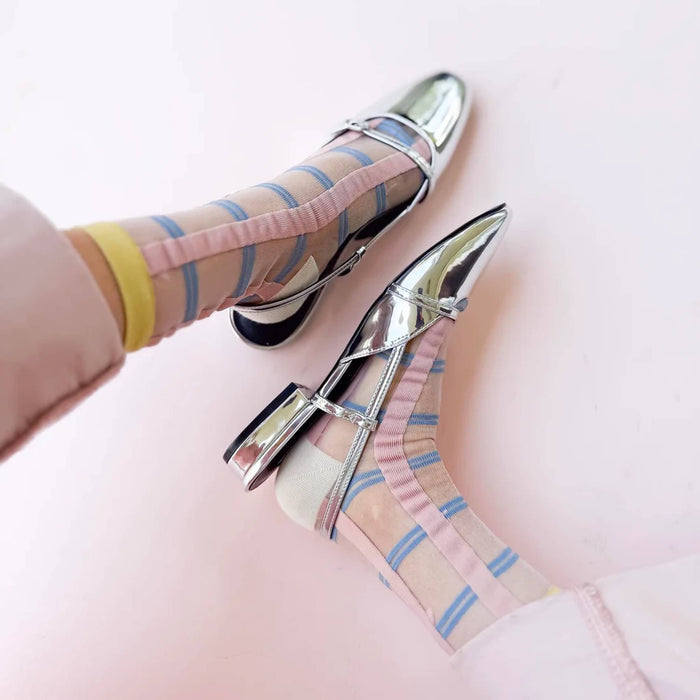 A pair of shiny silver pointed-toe flats with crisscross ankle straps, shown being worn with pastel pink, blue, and yellow striped socks on a soft pink background.