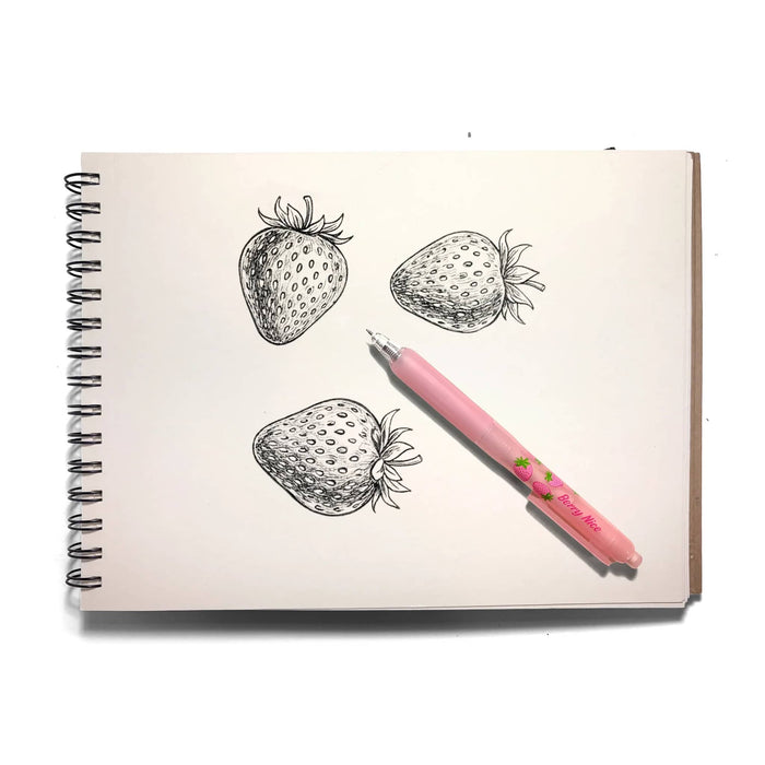 A pink mechanical pencil with a strawberry design lies diagonally across an open spiral-bound sketchbook. The sketchbook page features three hand-drawn black ink illustrations of strawberries on off-white paper, all photographed against a clean white background.