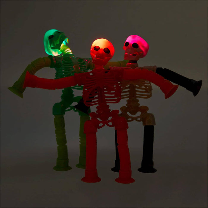 Three articulated plastic skeleton toys with glowing heads, posed side by side against a dark gray background. The left skeleton has a green glowing head and body, the center skeleton is red with a glowing orange head, and the right skeleton has a pink glowing head and black legs.