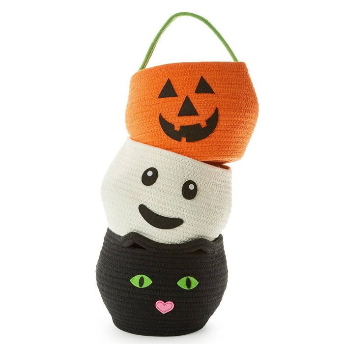 A Halloween-themed woven basket stack with three stacked baskets: an orange pumpkin basket on top with a green hanging loop, a white ghost face basket in the middle, and a black cat face basket on the bottom with green eyes and a pink heart nose.