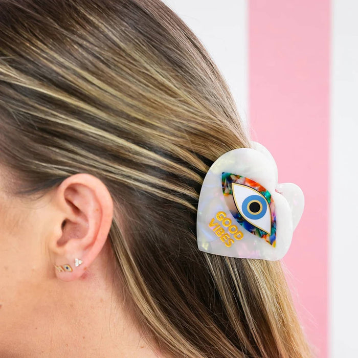 A close-up of a person's side profile showing a white iridescent heart-shaped hair clip with a colorful All-Seeing Eye design and the text 'GOOD VIBES' in gold, worn in light brown hair.