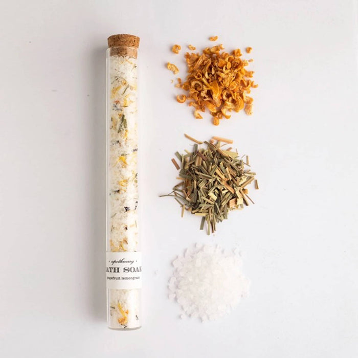 Glass tube with cork stopper filled with bath salt soak with piles of ingredients beside it, on a white background.