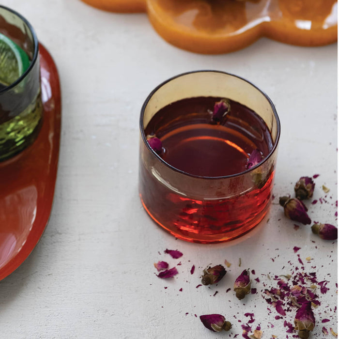 A hammered amber glass tumbler, filled with liquid and dried rosebuds, set on a white surface with scattered rose petals and dried flower pieces.